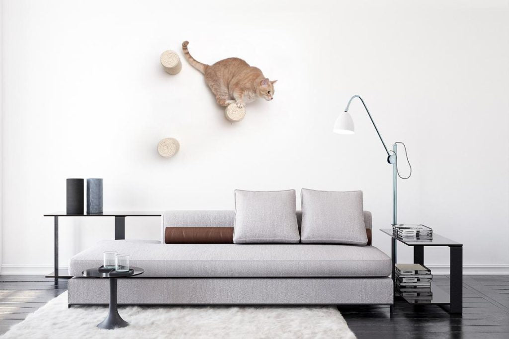 Photo of large orange cat atop 1 of 3 floating sisal poles, mounted above a low sofa in a neutral-toned living space.